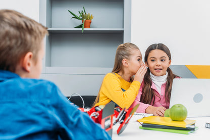 Two children whispering to each other at a table with a third child looking on.