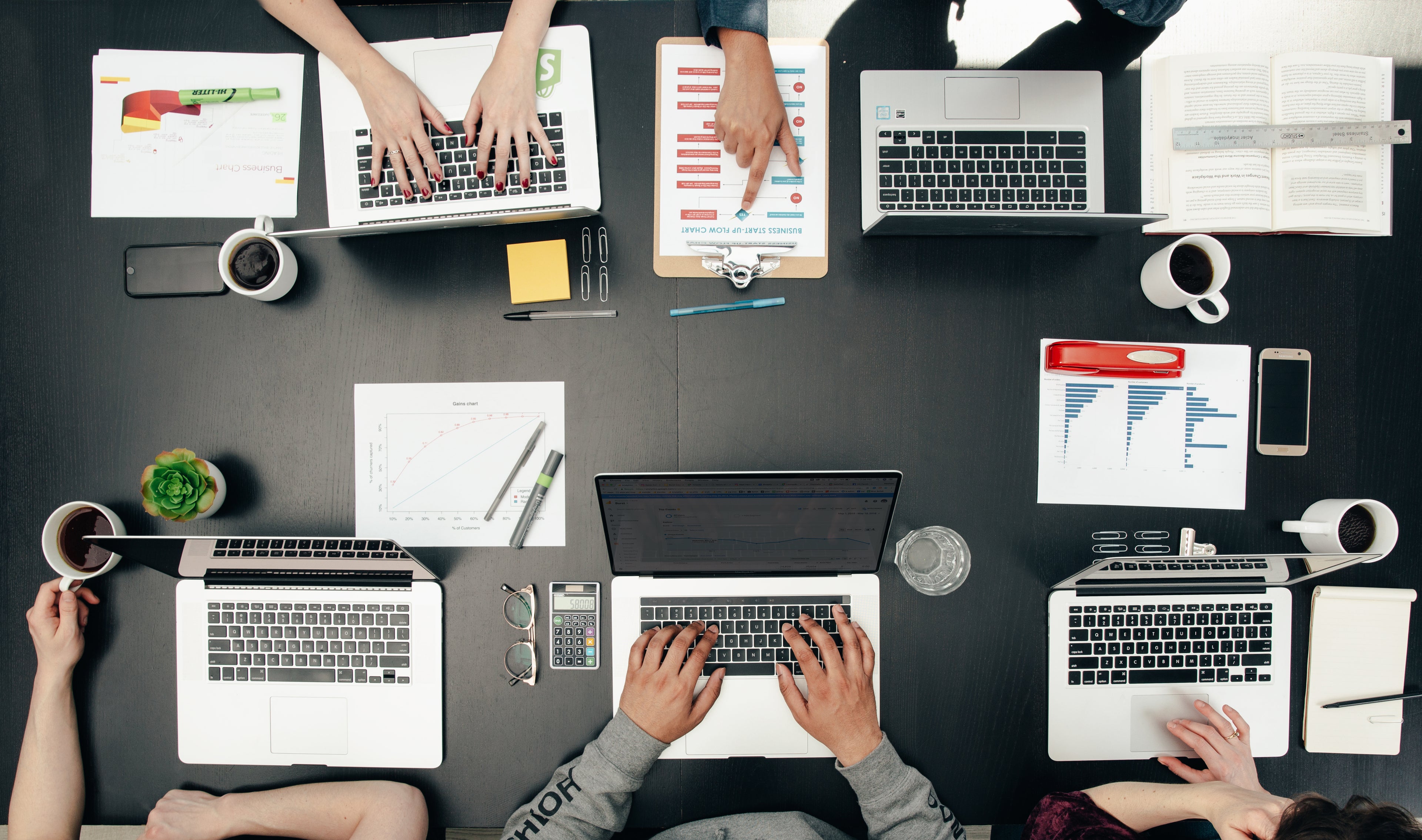 Top-down view of a busy office desk with multiple laptops, papers, and people working.